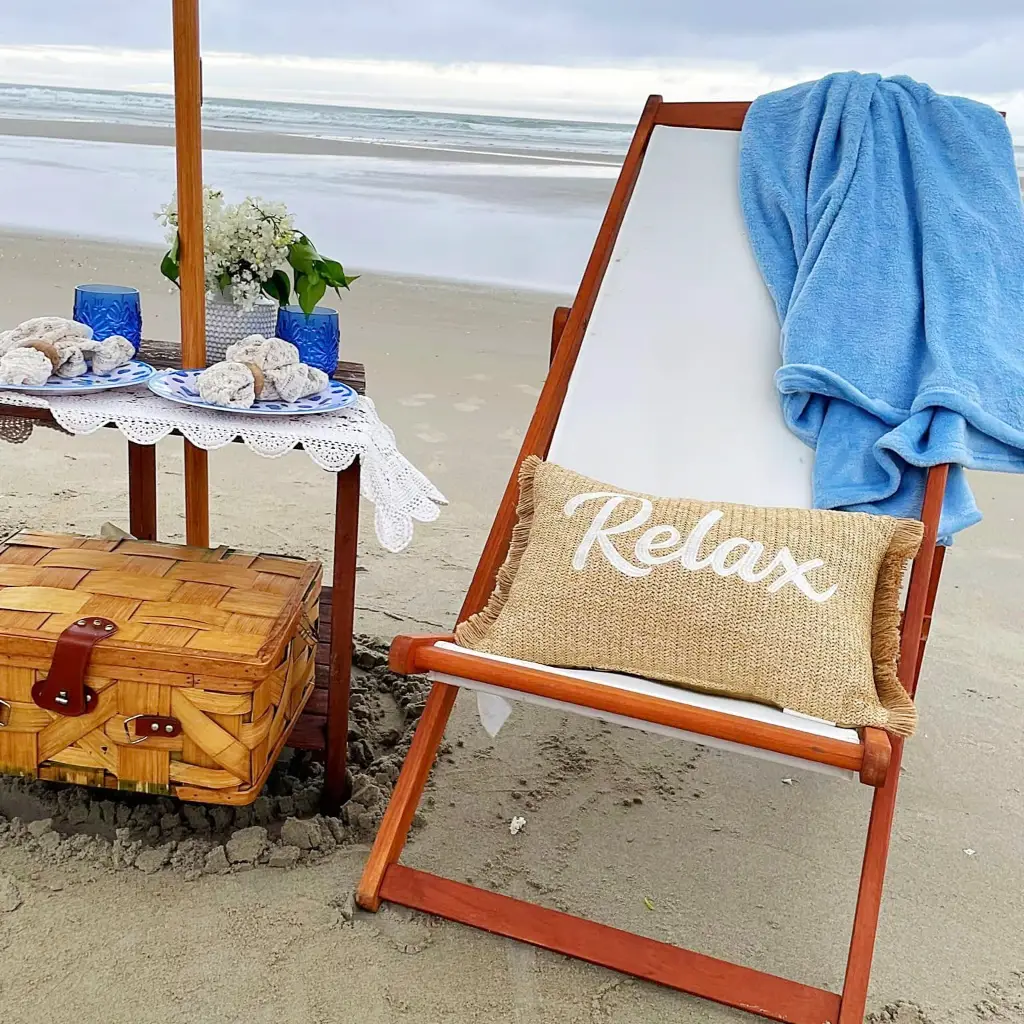 A small table setup on the beach with plates and a picnic basket along with a chair with a pillow that says 'Relax'.